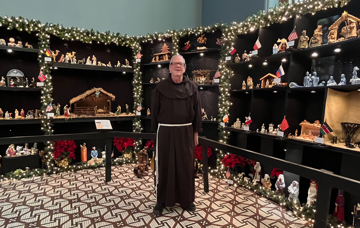 A Franciscan friar stands in front of a large display of Christmas crèches