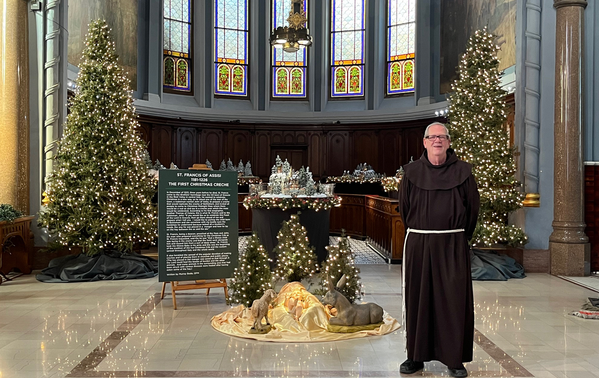 A friar stands in a Church next to a life-size nativity scene surrounded by Christmas trees.