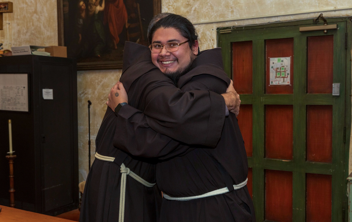 A friar shares a congratulatory hug with one of the newly-vested novices. The novice is grinning widely.