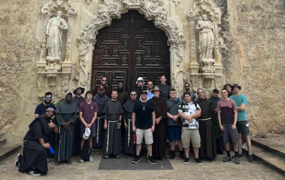 A group of 23 friars and men in discernment stand in front of the bronze doors of an old mission church.