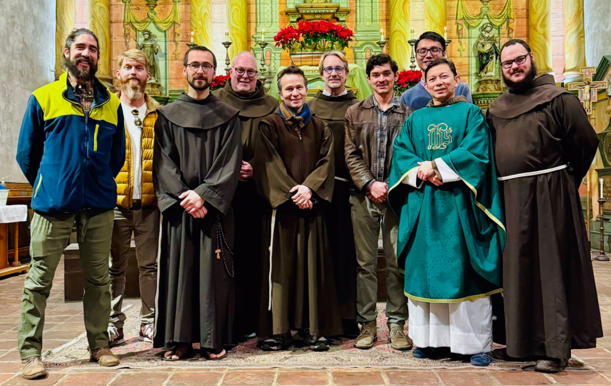 A group of men, some wearing Franciscan habits, stand in front of the altar of a historic mission church