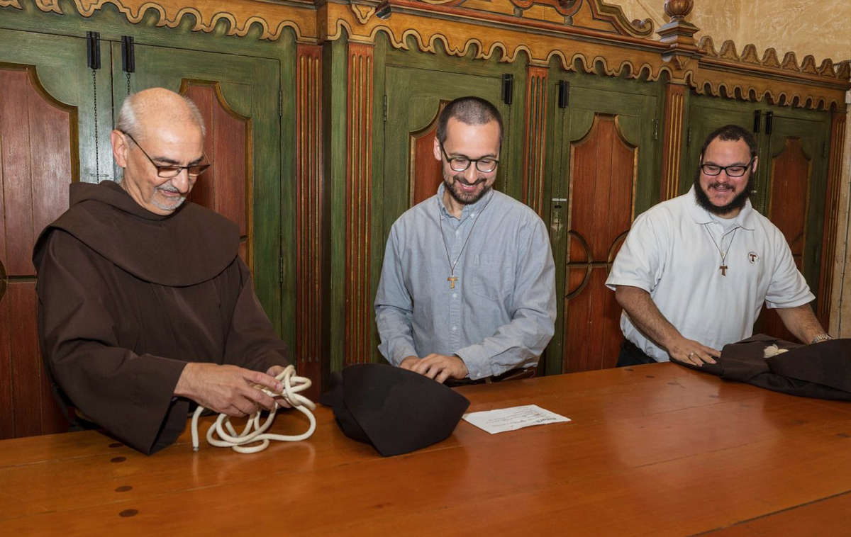 Two smiling men watch as an older friar unfolds a Franciscan habit.