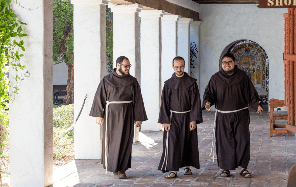 Three men dressed in the brown Franciscan habit walk in the cloister of an old mission-style building