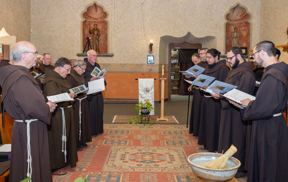 A group of 15 Franciscan friars pray together in a mission-style chapel.