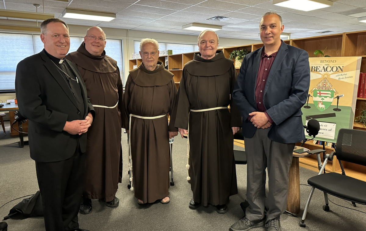 Five men, one dressed as clergy, three wearing friar habits, and one in a sports coat, standing in a room.