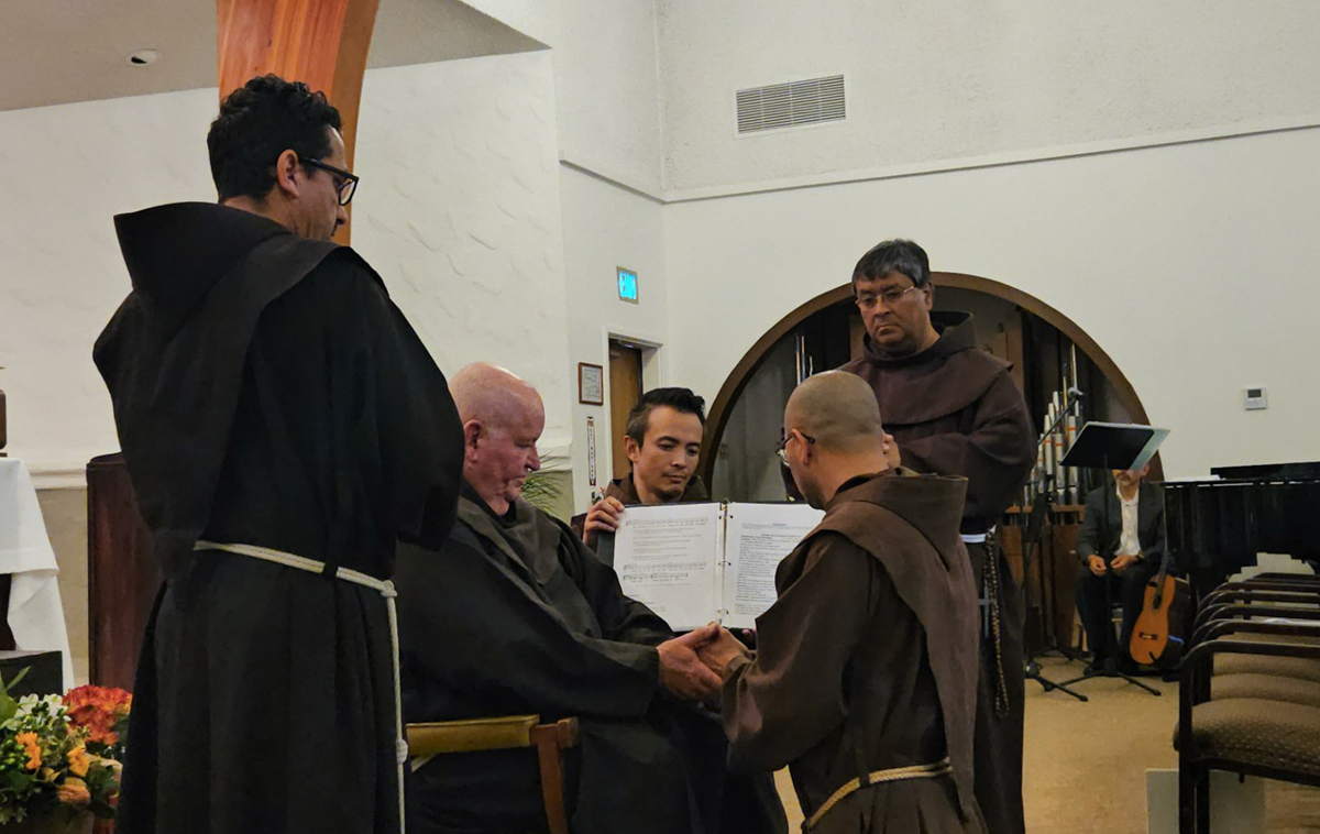 Br. Martin kneels before another friar, who is holding his hands, while other friars look on.