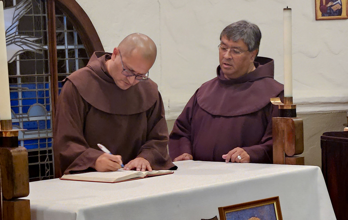 A friar signs his name in a book while another friar looks on.