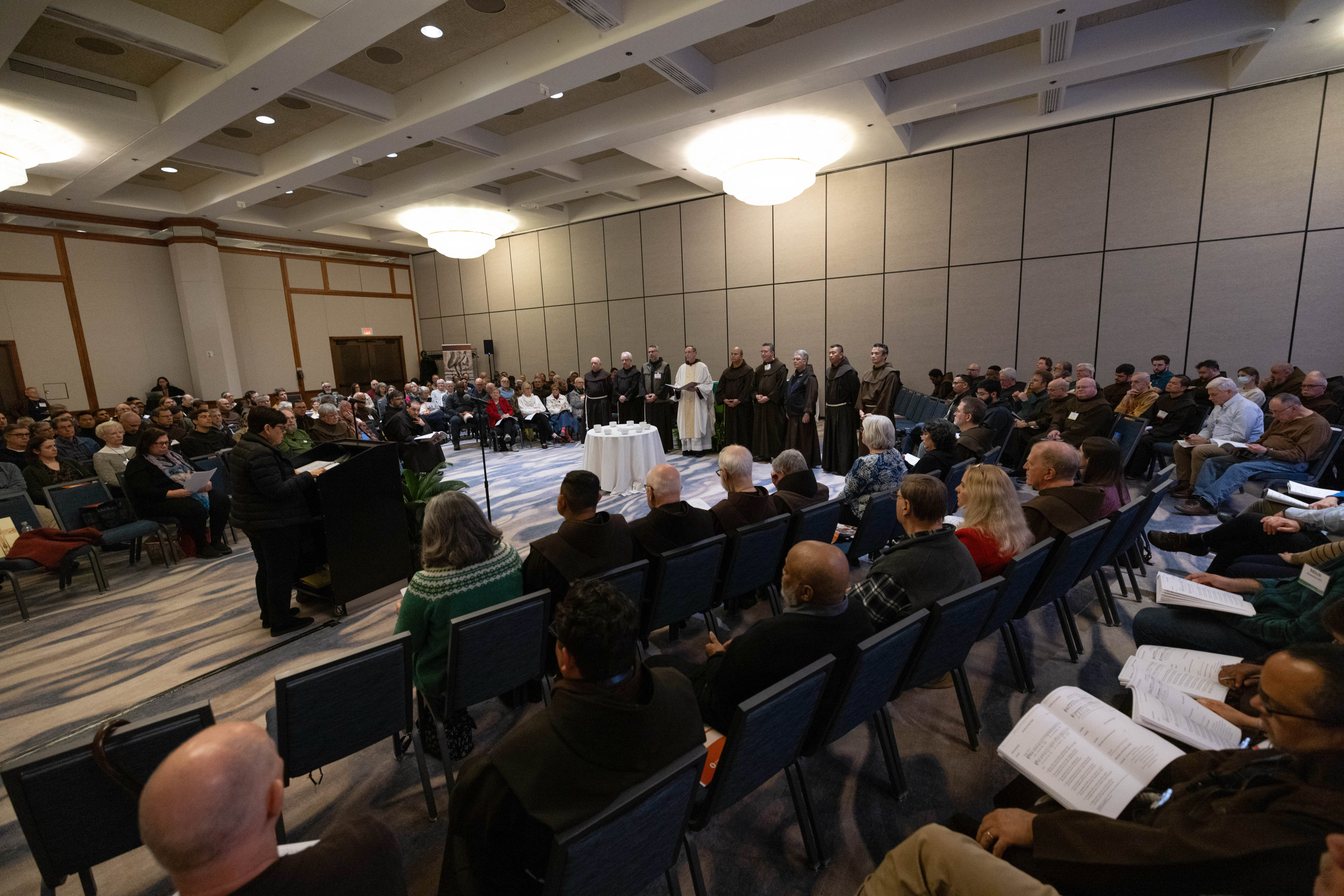 Dozens of people sit in chairs on opposite sides of a room with a podium and table in between. Nine friars stand by a wall behind the table.