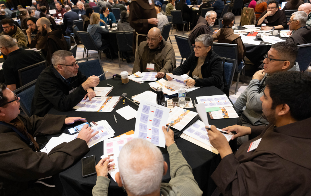 Seven people sit around a table with papers, pens and drink containers in front of them. One is pointing at another with an ink pen.