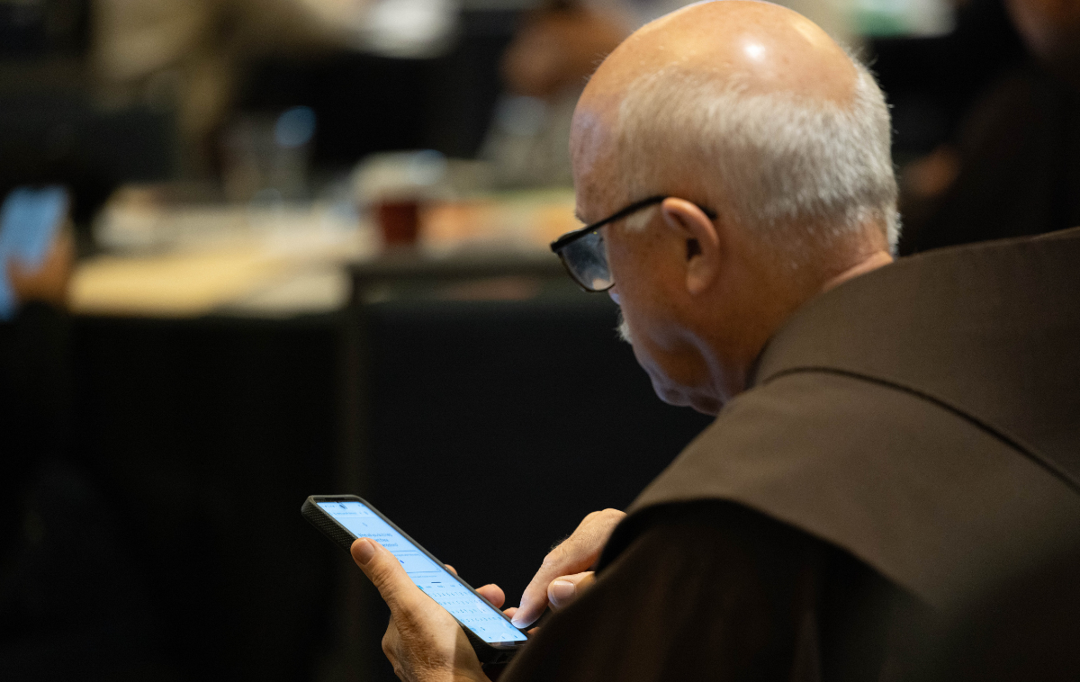 A man wearing eyeglasses and a friar habit reads from the screen of a mobile device.