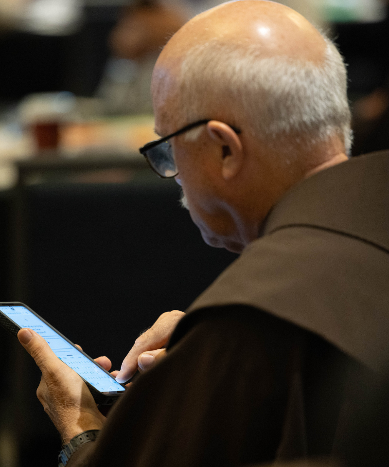 A man wearing a friar habit and eyeglasses looks at a smartphone screen.