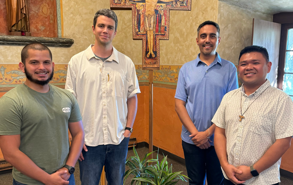 Four men stand in a room beside a plant and in front of a crucifix. There is a statue of Jesus Christ behind them.