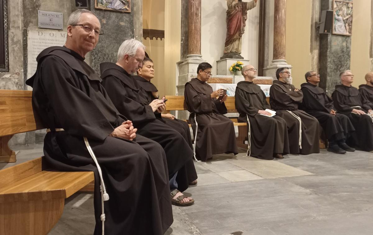 Nine men in friars habits sit on pews in a church.