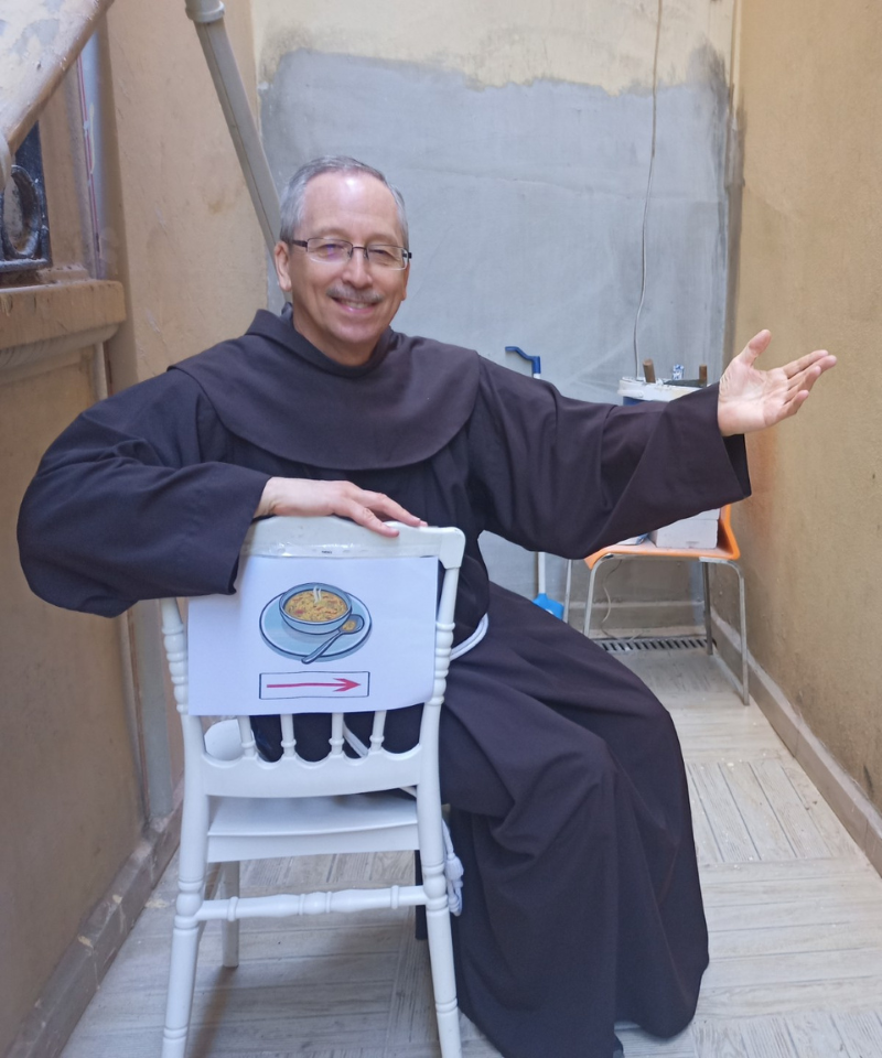 A smiling man wearing glasses and a habit sits on a white chair. A sign on the chair points to the entrance of a soup kitchen.