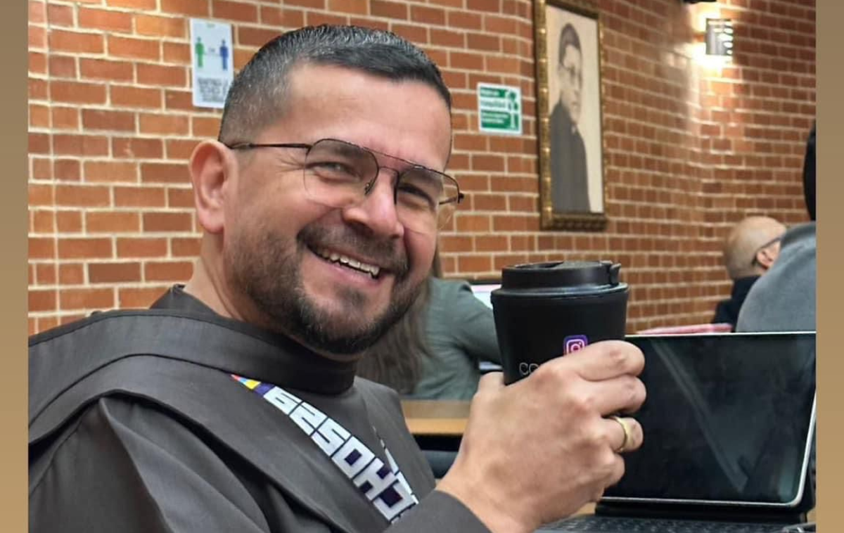 A bearded, smiling man is wearing glasses, a friar habit, and a lanyard. He is sitting in a brick-walled building, sitting in front of a computer, and holding a coffee cup.