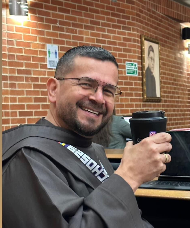 A bearded, smiling man wearing glasses, a friar habit, and a lanyard holds a coffee cup. He is sitting in a brick-walled room in front of a computer.