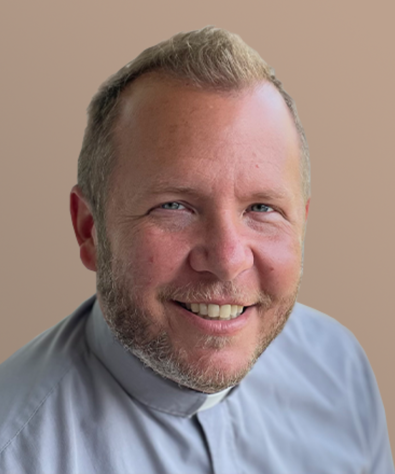 A man wearing a blue shirt and clergy collar smiles.