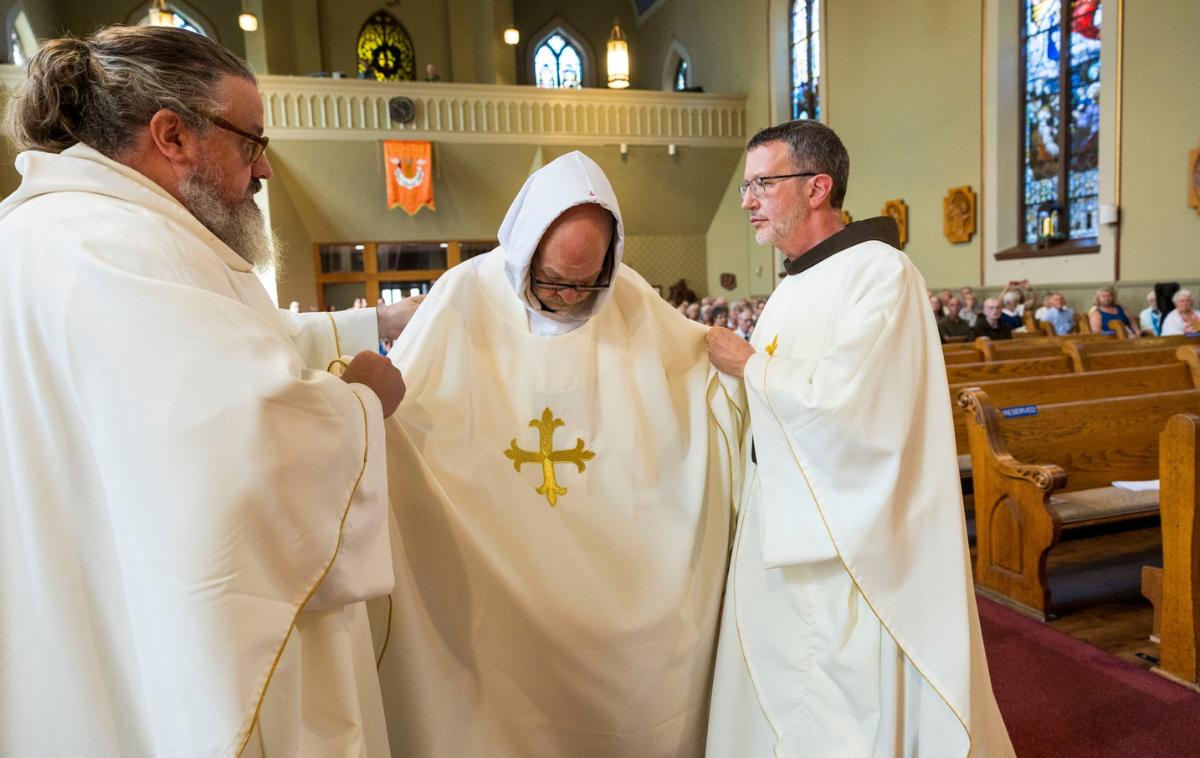 Two men place vestments on another man in a Catholic Church.