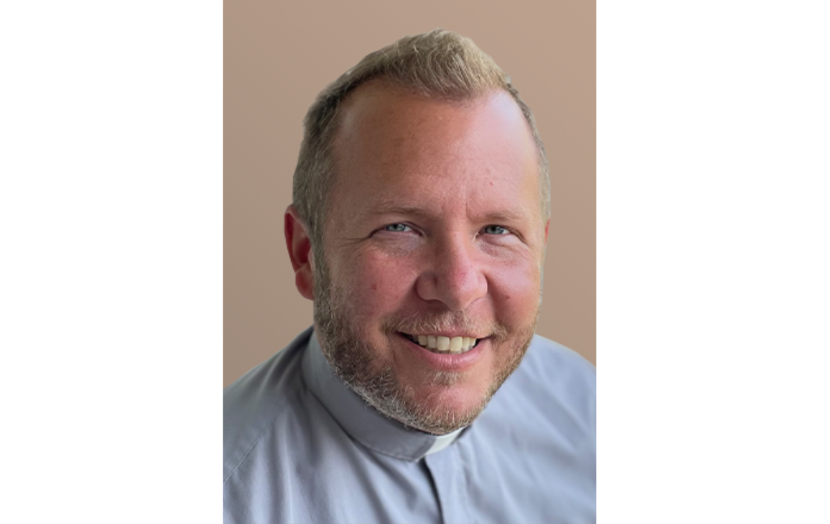 A smiling man in a blue shirt and clergy collar poses for a photo.