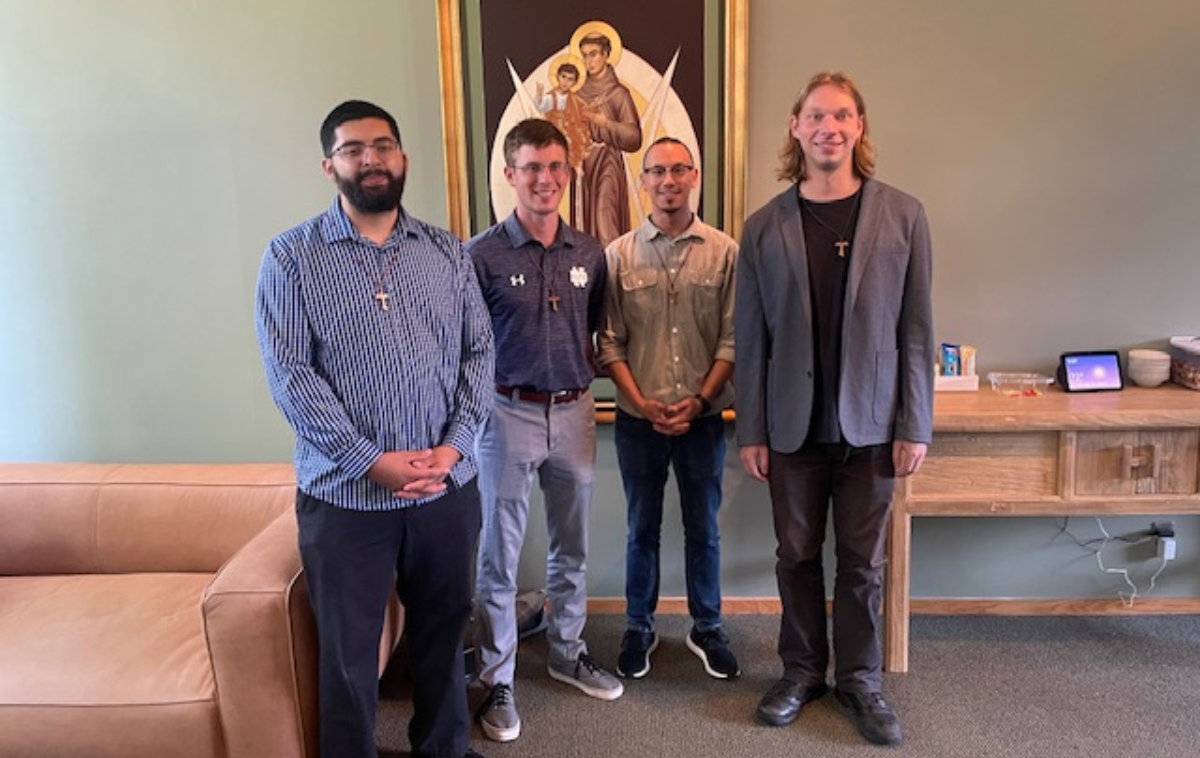 Four men stand in a room between a couch and a table and in front of an image of a saint.