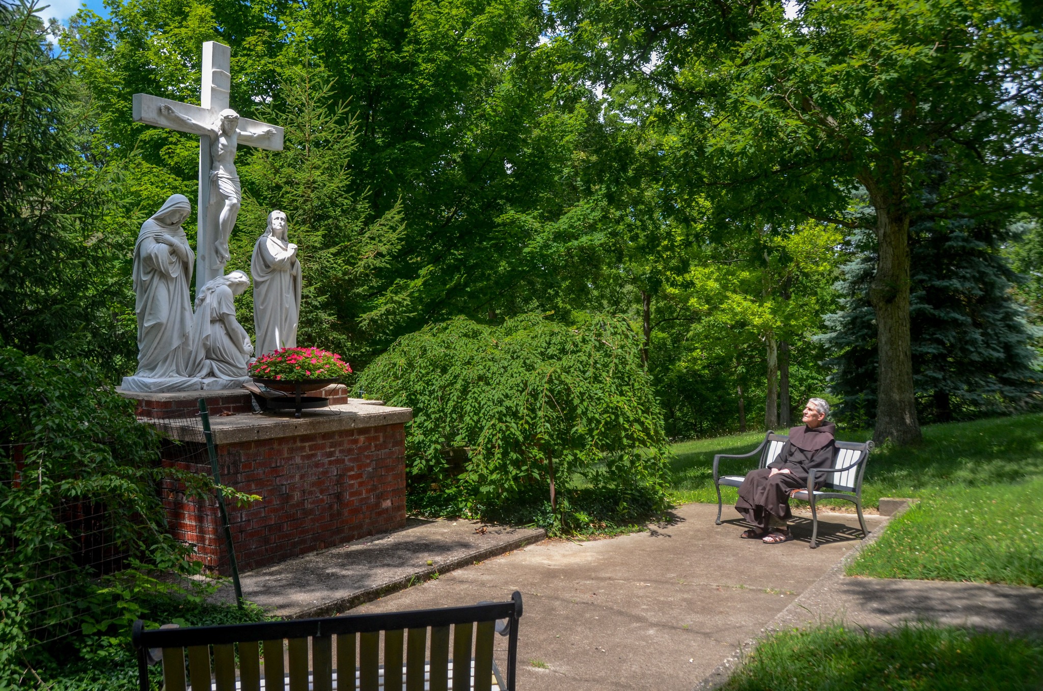 A man in a friar habit sits on a bench in a sanctuary looking at a statue of Jesus Christ on a cross with two women and a man depicted in front of him. There is a container of flowers in the foreground with a second bench on a paved surface.