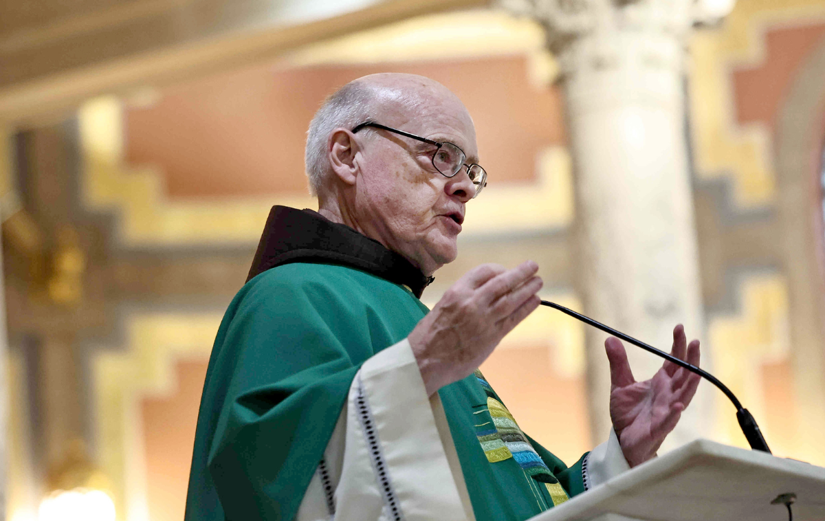 A priest preaches at the pulpit, gesturing toward the ceiling with his hands