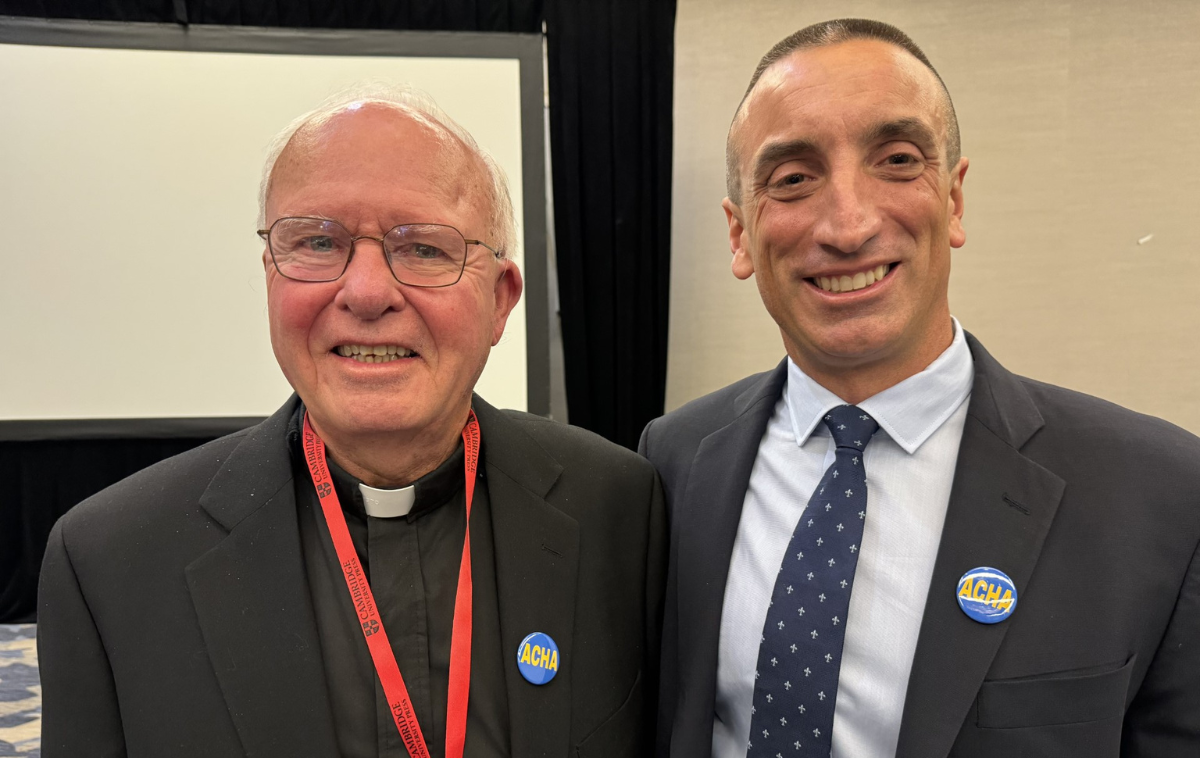 Two men smile at the camera. One is wearing black clothes and a priest's collar. The other is wearing a suit and tie. Both are wearing small button pins with ACHA in big letters.