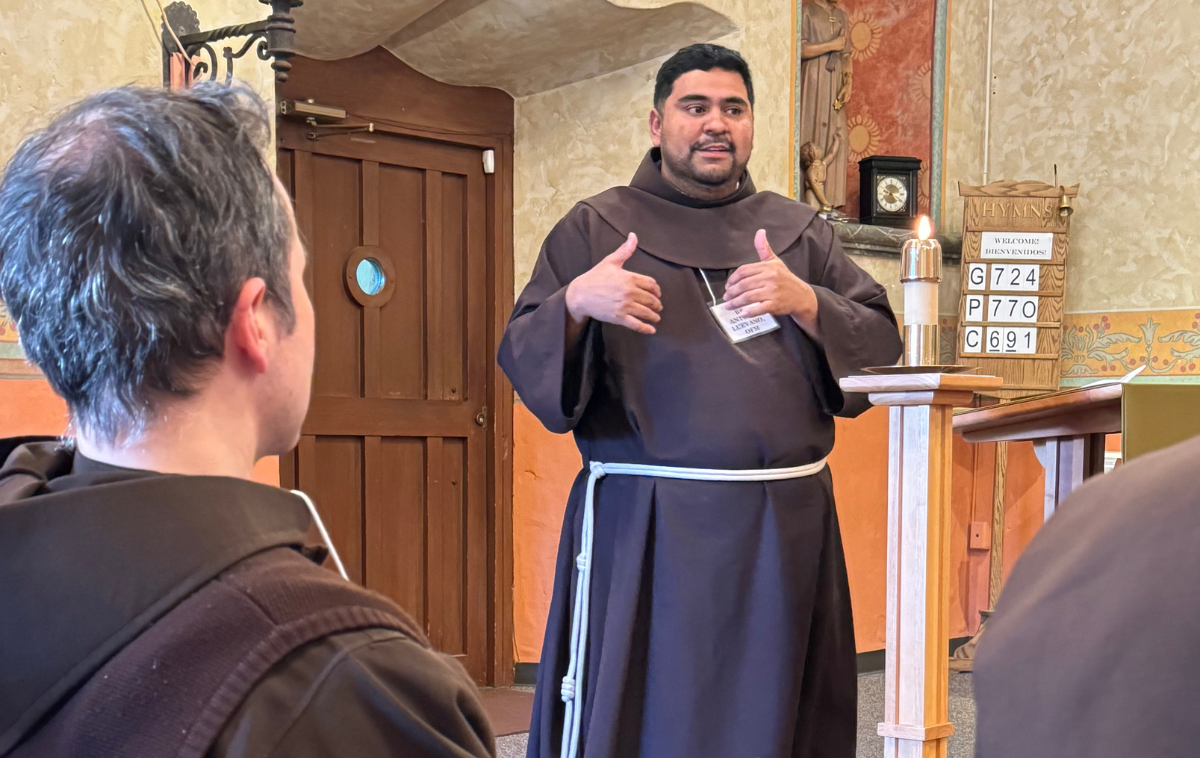 A Hispanic friar in his 30s speaks in front of a small group in a historic mission church