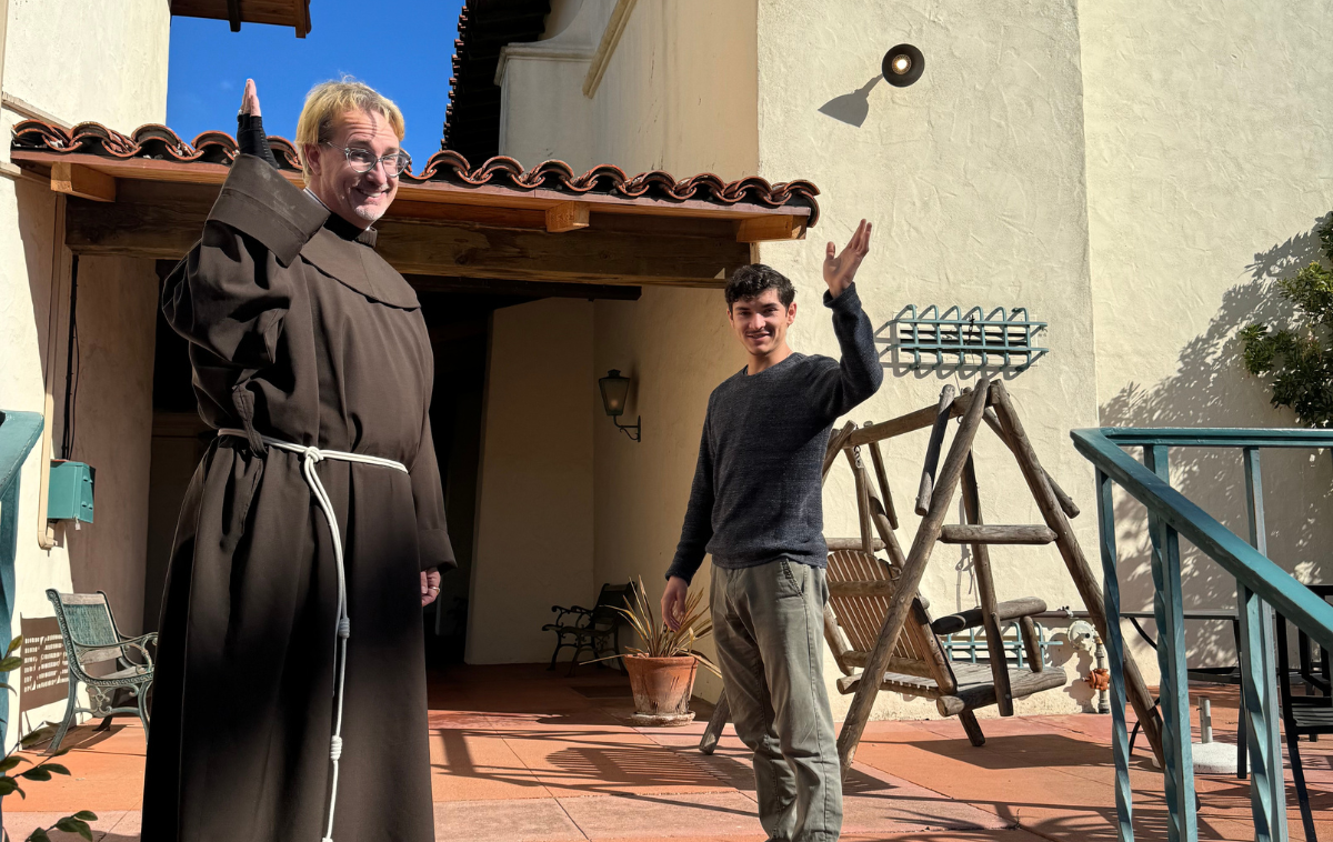 A friar and a man in his 20s smile and wave at the camera