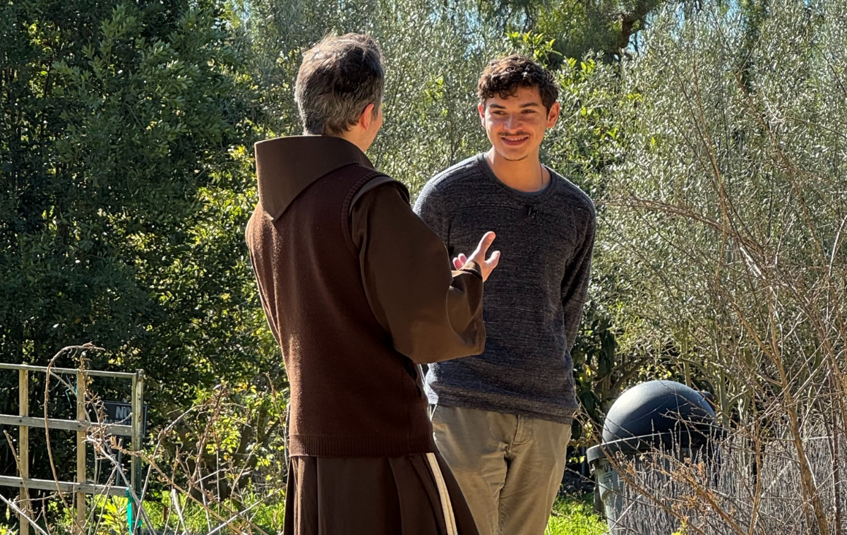A friar speaks with a man in his 20s who is smiling