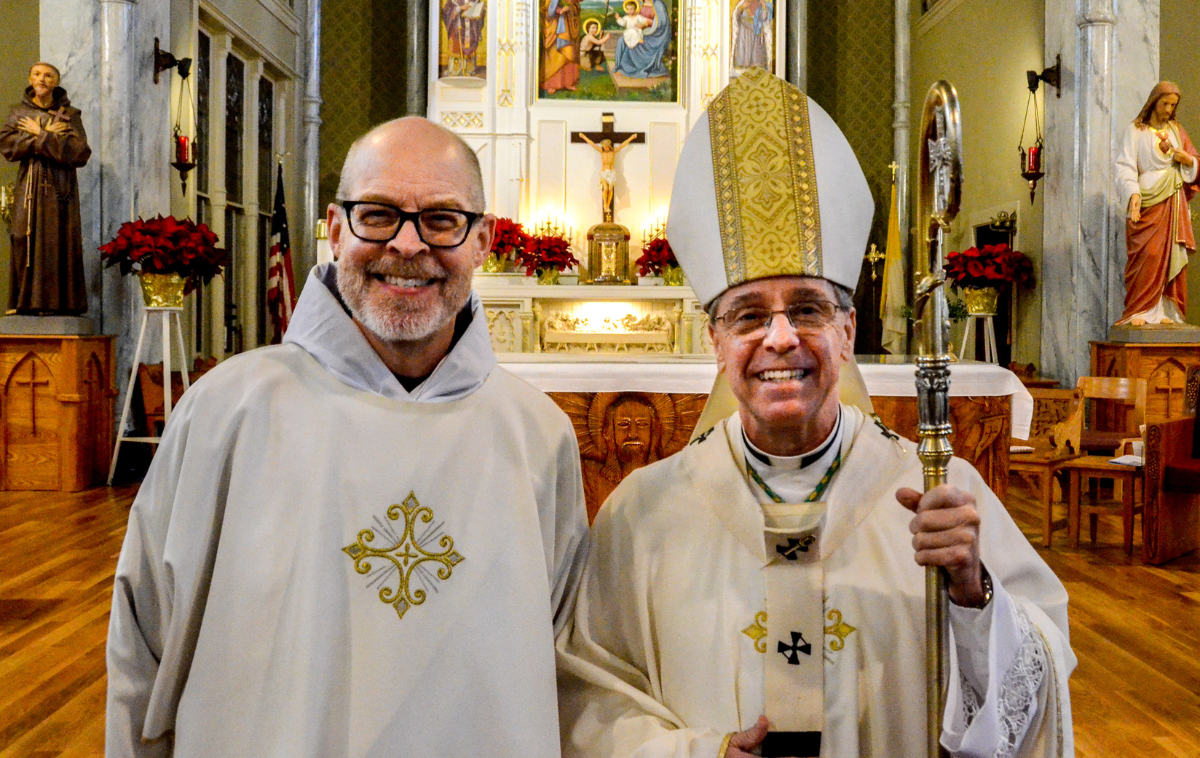 A deacon and an archbishop stand smiling in front of an altar in a church