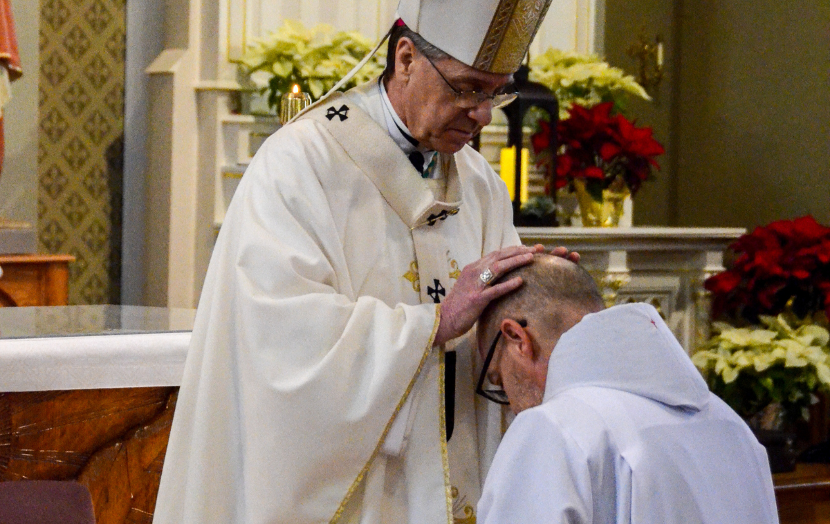 A bishop rests his hands in prayer on the head of a deacon kneeling in front of him