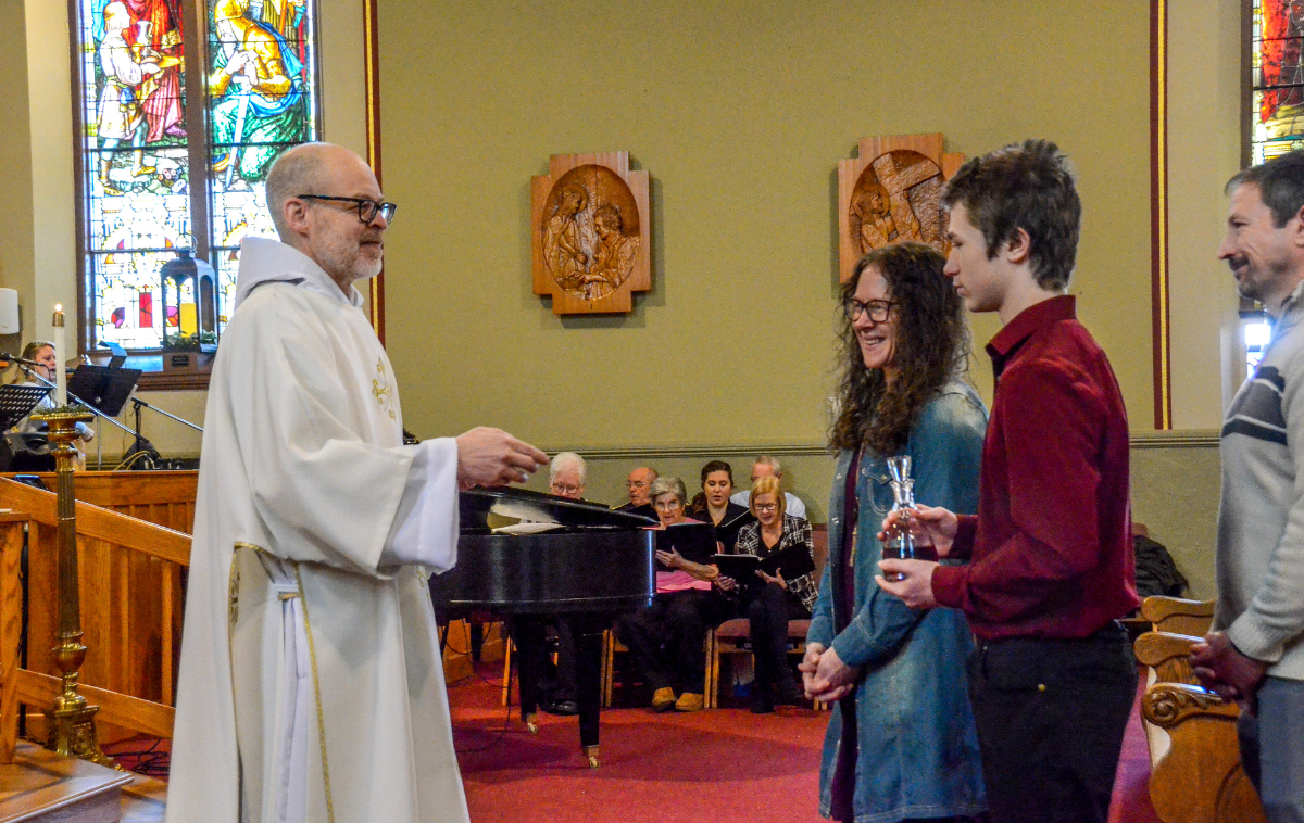 A deacon smiles as he accepts the presentation of the gifts from a young woman and her son