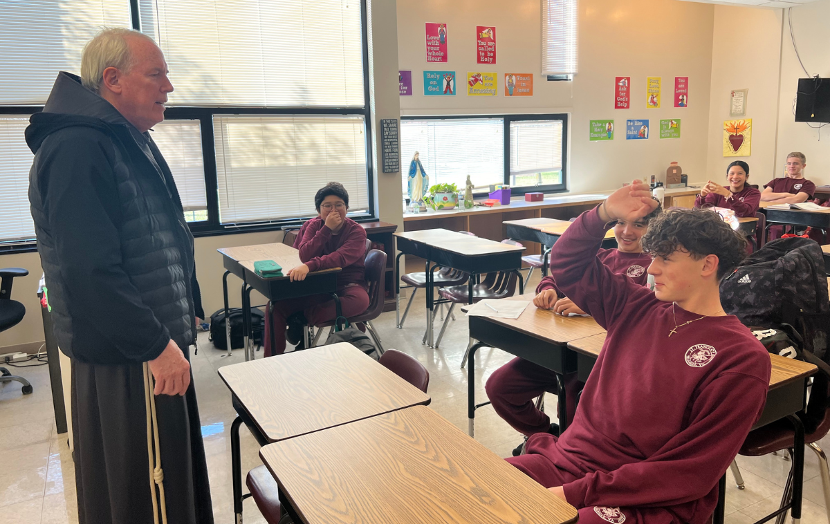 A friar stands at the front of a classroom and addresses a student raising their hand