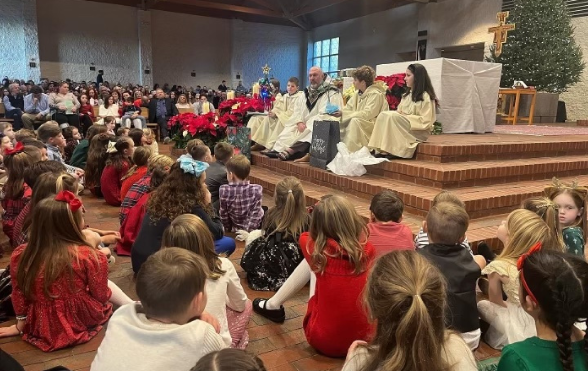 A priest sits on the steps of the altar and speaks to a group of elementary school students