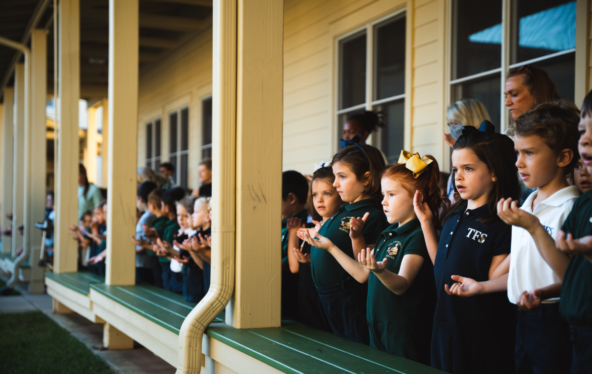 Elementary school students hold their hands up in prayer as they stand in a school courtyard