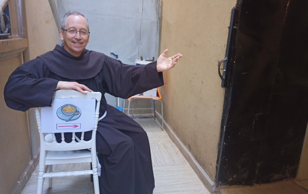 A smiling man in glasses and a friar habit sits in a white chair with a sign on it pointing to the entrance of a soup kitchen