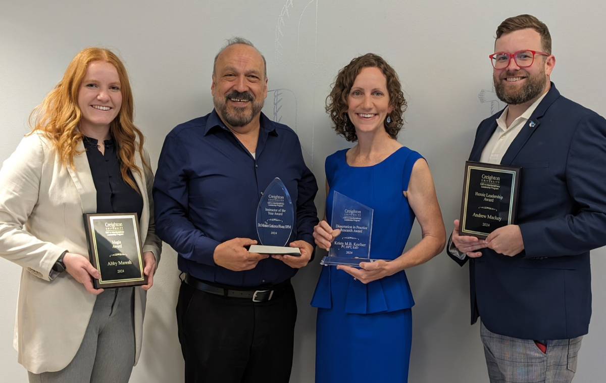 Moises stands among three other faculty members who received awards from Creighton University. He is smiling, wearing a dark blue shirt and holding a glass award with his name on it.