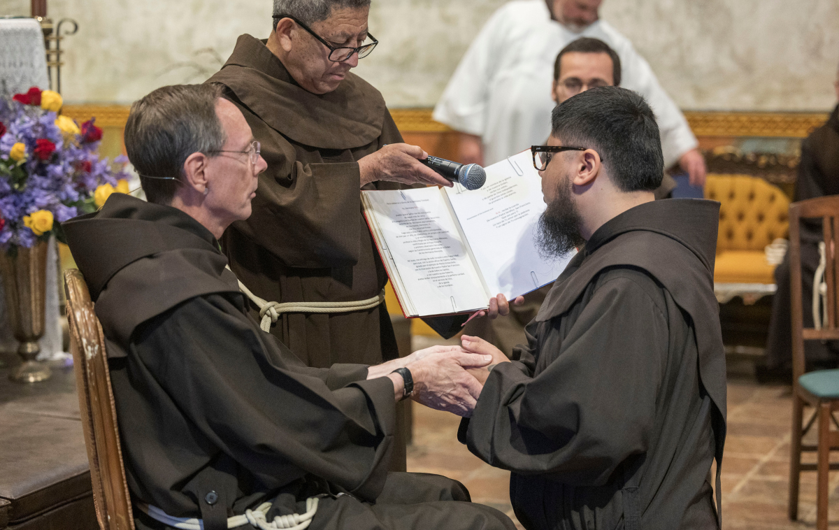 A young friar kneels in front of an older friar, who clasps his hands. The kneeling friar reads from a book held open before him.