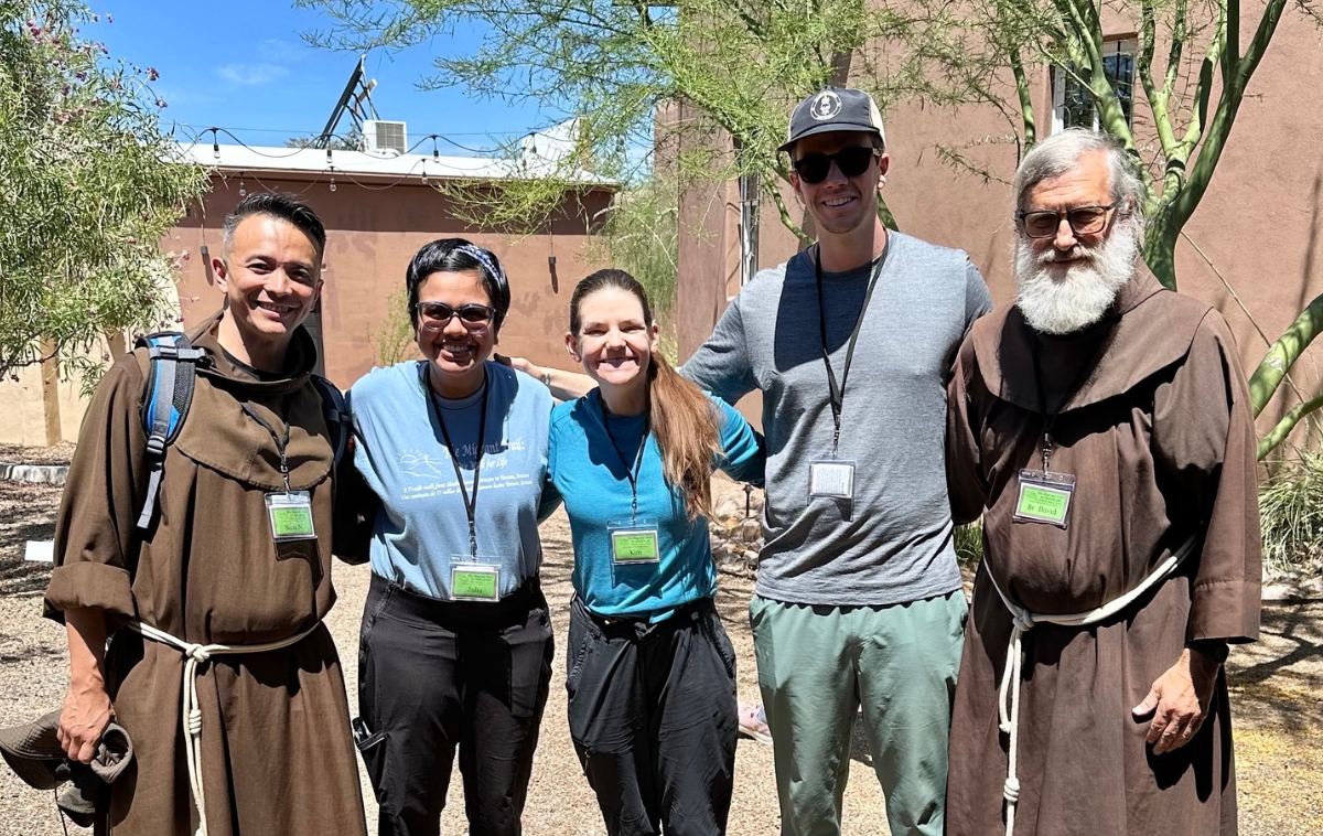 Five people wearing lanyards with green badges smile at the camera. Two of them are Franciscan friars who are wearing their habits.