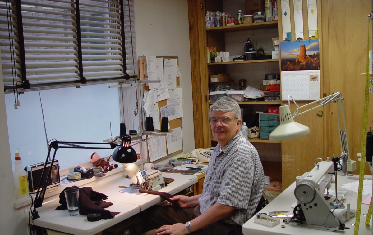 A man in his 60s wearing glasses and a white and blue plaid collared T-shirt sits smiling in front of a sewing machine. He is holding brown fabric in his hands.