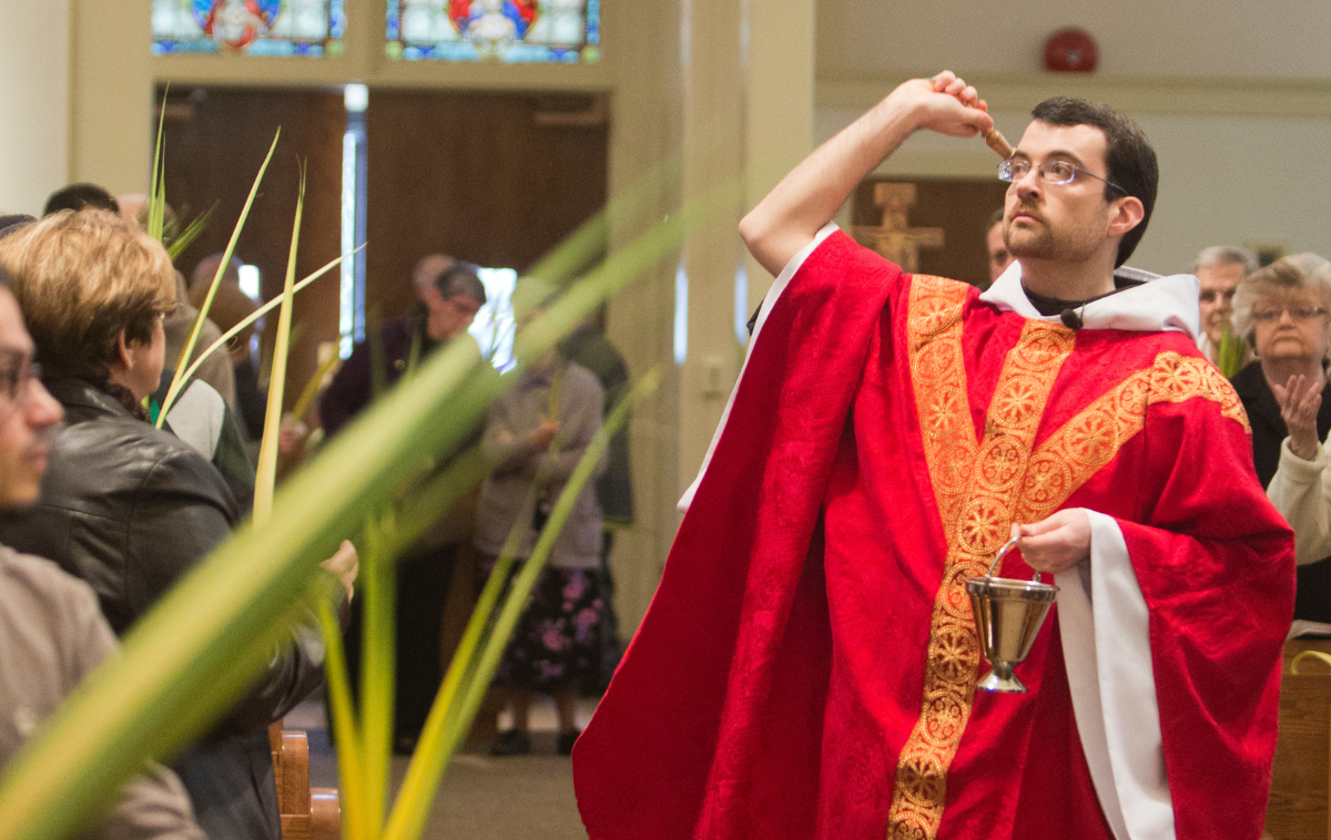 A priest wearing red vestments sprinkles holy water on a crowd of people holding palms
