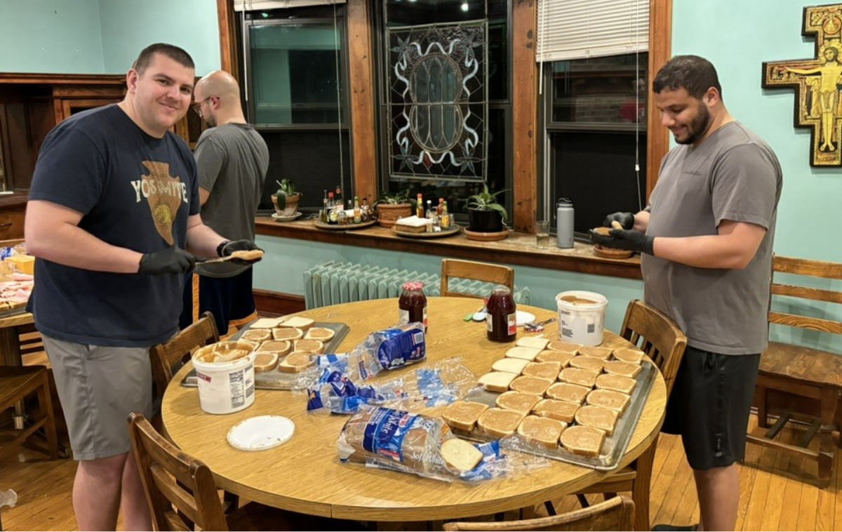 Three men in their 20s and 30s smile as they make peanut butter and jelly sandwiches