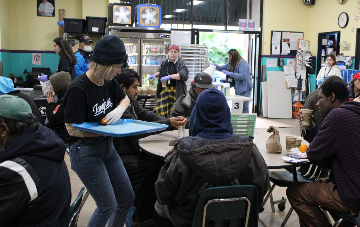 A person with shoulder-length blonde hair and a black beanie hat serves food to people sitting at a round table in a dining room