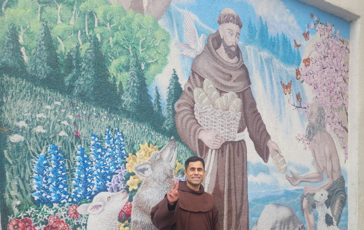 A friar holds up a peace sign in front of a beautiful, intricate mural of St. Francis sharing bread with a leper as forest creatures watch.