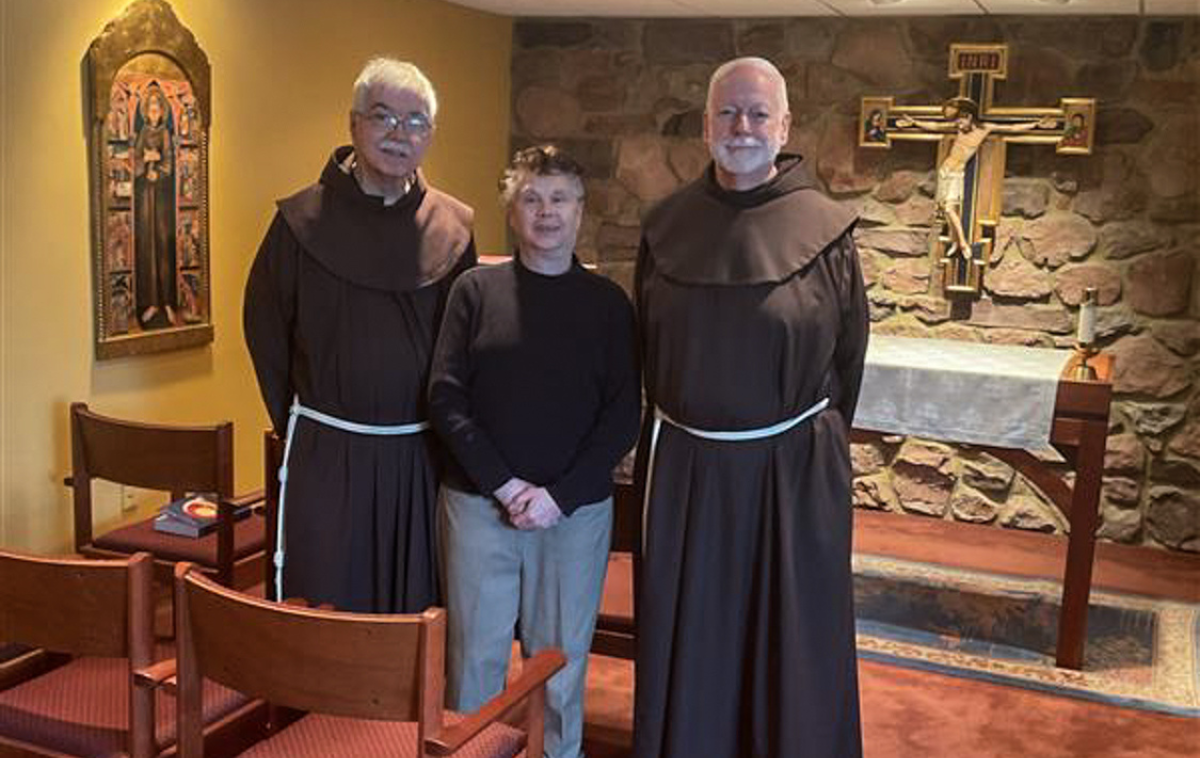 A woman stands between two Franciscan friars in front of an altar and the San Damiano Cross in a chapel