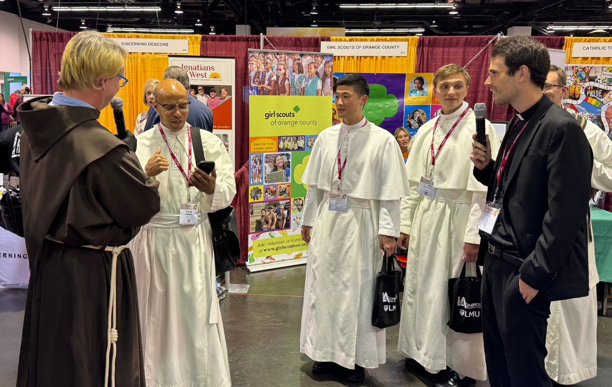 A friar wearing a brown robe talks with three men in white robes and a priest wearing black clothing and a white collar