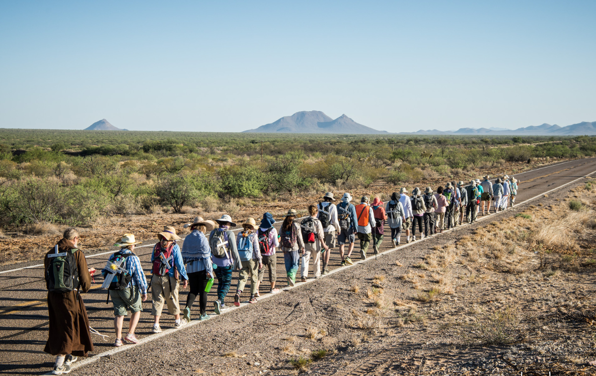 A line of 30 people walks alongside a road in a rural part of the Arizona desert. There are no buildings or cars in sight. There are mountains in the far distance on the horizon.
