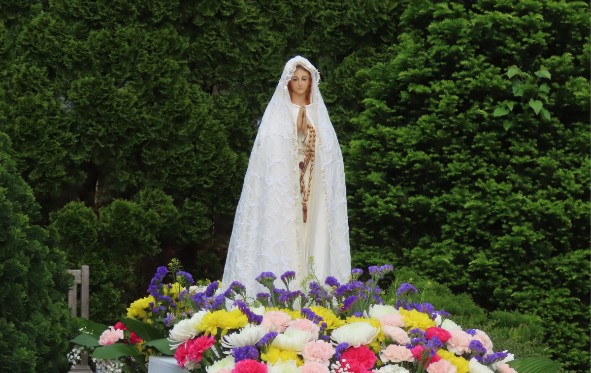 A statute of Mary wearing a beautiful white lace veil stands in the center of a colorful floral arrangement made of white, pink, red, yellow and purple flowers