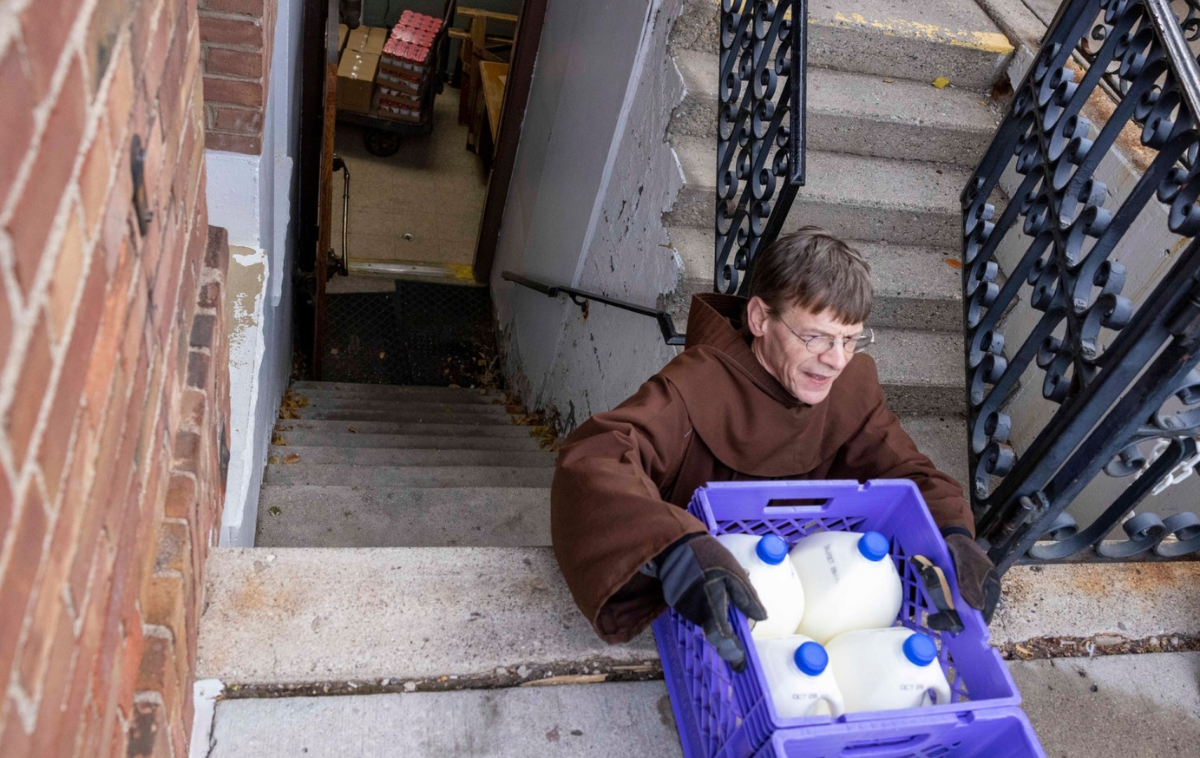 A friar lifts a crate containing four gallons of milk at the top of a set of stairs.